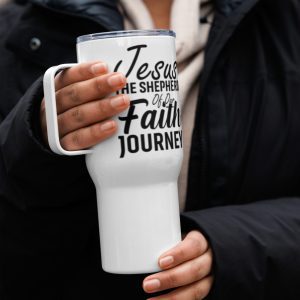 A person in a dark coat holds a white Jesus The Shepherd Of Our Faith Journey Travel Mug with a large handle. The mug has the text "Jesus, the Shepherd of your Faith Journey" printed in bold, black script. The background is blurred, focusing attention on the mug and the person's hands. christian gifts, religious gifts, gifts of faith, faith based gifts, faith inspired gifts