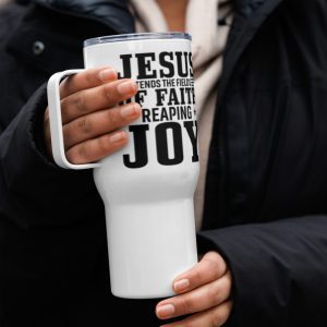 A person wearing a black jacket and a beige scarf holds a white travel mug with bold black text that reads "JESUS TENDS THE FIELDS OF FAITH REAPING JOY." The background is slightly out of focus, highlighting the mug and the person's hands. christian gifts, religious gifts, gifts of faith, faith based gifts, faith inspired gifts