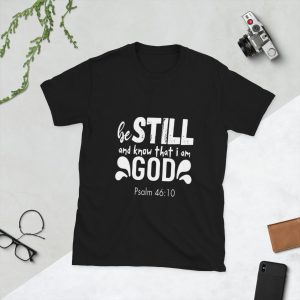 A Be Still and Say that I Am God "Psalm 46:10" Unisex T-Shirt featuring white text on a black background is laid flat on a white surface, surrounded by green leaves, a camera, glasses, and a notebook. christian gifts, religious gifts, gifts of faith, faith based gifts, faith inspired gifts