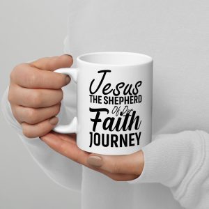 A pair of hands hold the "Jesus The Shepherd Of Our Faith Journey Coffee Cup" by Judeo Gifts, a white mug adorned with black text that reads, "Jesus, the shepherd of our faith journey." The person wearing a white long-sleeve shirt demonstrates the perfect blend of sentiment and simplicity. Only their hands are visible in the image. christian gifts, religious gifts, gifts of faith, faith based gifts, faith inspired gifts