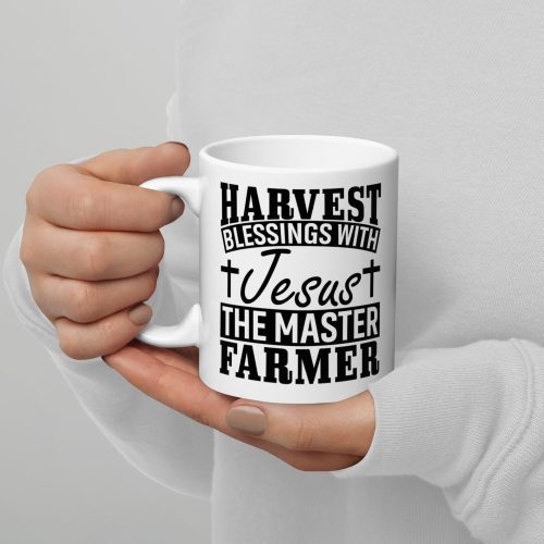 A person wearing a white sweater holds the Harvest Blessing With Jesus The Master Farmer Coffee Cup from Judeo Gifts, adorned with the message, "HARVEST BLESSINGS WITH Jesus THE MASTER FARMER." The text is in bold black lettering and is surrounded by small crosses around the word "Jesus. christian gifts, religious gifts, gifts of faith, faith based gifts, faith inspired gifts