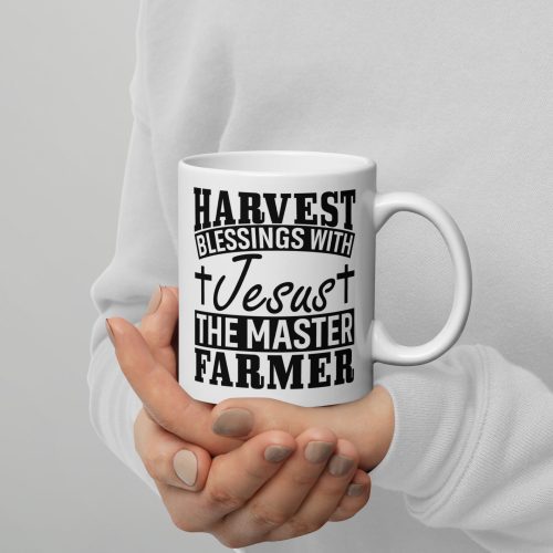 A person wearing a white sweater holds the Harvest Blessing With Jesus The Master Farmer Coffee Cup—a white mug featuring black text that reads, "Harvest blessings with Jesus the Master Farmer," along with two small cross symbols. This beautiful Christian gift spreads the joy and love of faith. christian gifts, religious gifts, gifts of faith, faith based gifts, faith inspired gifts