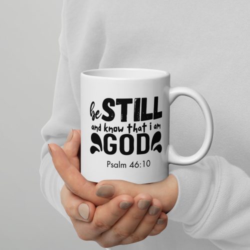 A person wearing a white sweater holds a "Be Still and Say that I Am God 'Psalm 46:10'" Coffee Cup, adorned with decorative elements around the black text. Only the hands and torso of the person are visible, showcasing one of many faithful gifts perfect for any believer. christian gifts, religious gifts, gifts of faith, faith based gifts, faith inspired gifts