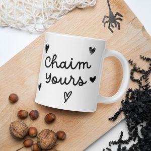 A white glossy mug called "Chaim Yours - Jewish Saying Meaning to Life!" features the text "Chaim Yours" in black cursive, along with small black heart illustrations. The mug is set on a wooden surface adorned with scattered hazelnuts, black decorative shreds, and a spider decoration nearby—an ideal addition to your collection of Faithful Gifts. christian gifts, religious gifts, gifts of faith, faith based gifts, faith inspired gifts