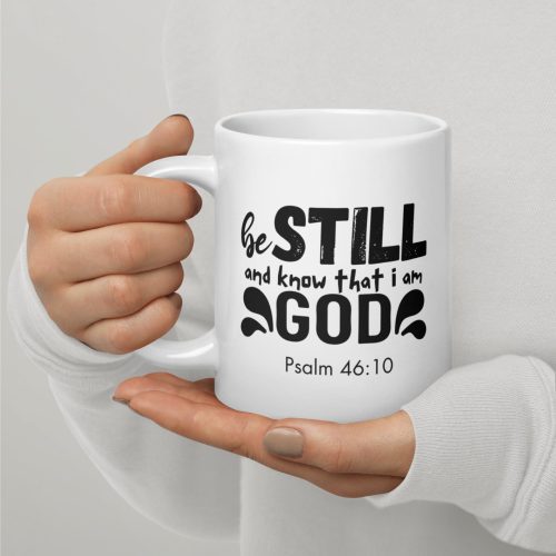 A person holds the "Be Still and Say that I Am God 'Psalm 46:10'" Coffee Cup, featuring text in bold black font. The mug stands out against a plain background while the individual's nails are polished in a neutral color, and they are wearing a light-colored, long-sleeved top—making it an ideal choice among religious gifts. christian gifts, religious gifts, gifts of faith, faith based gifts, faith inspired gifts