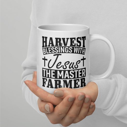 A person wearing a light-colored sweater is holding a "Harvest Blessing With Jesus The Master Farmer Coffee Cup," which features black text that reads "HARVEST BLESSINGS WITH Jesus THE MASTER FARMER" and two small cross graphics on either side of the word "Jesus." This makes an ideal present for those seeking faithful gifts. christian gifts, religious gifts, gifts of faith, faith based gifts, faith inspired gifts