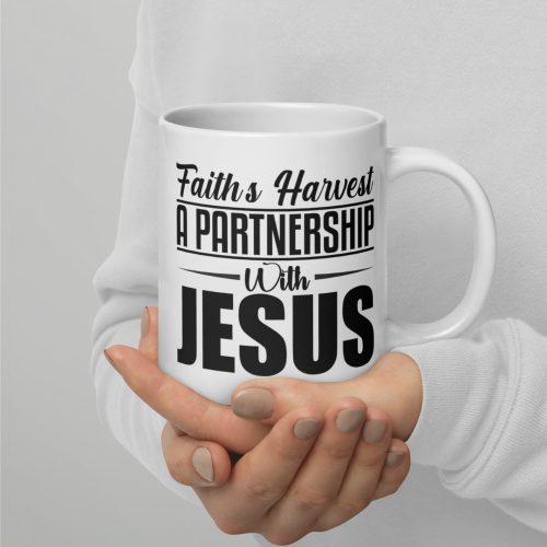 A person is holding a Faiths Harvest A Partnership With Jesus Coffee Cup with both hands. The mug, available from Judeo Gifts, features the text "Faith's Harvest A Partnership With Jesus." The background is a plain light grey, and the individual is wearing a white long-sleeve shirt. christian gifts, religious gifts, gifts of faith, faith based gifts, faith inspired gifts