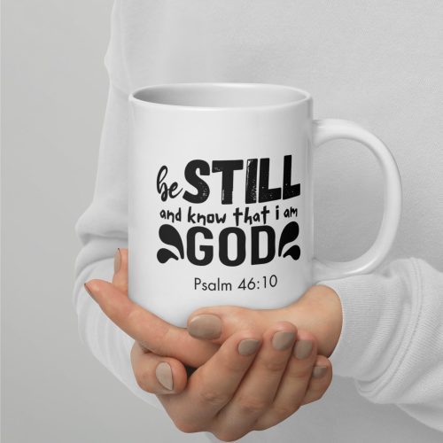 A person wearing a white long-sleeve shirt holds the "Be Still and Say that I Am God 'Psalm 46:10' Coffee Cup," a white ceramic mug with the phrase "Be STILL and know that I am GOD" and "Psalm 46:10" written on it in black letters. The individual's nails are painted a neutral color, creating an ideal moment for showcasing Christian gifts of faith. christian gifts, religious gifts, gifts of faith, faith based gifts, faith inspired gifts