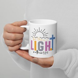 A person holds the "Be The Light Coffee Cup," featuring the text "be the LIGHT" and "Matthew 5:14," with a sun illustration above and a blue cross accompanied by a small yellow heart below. The person is dressed in a white long-sleeve top. christian gifts, religious gifts, gifts of faith, faith based gifts, faith inspired gifts