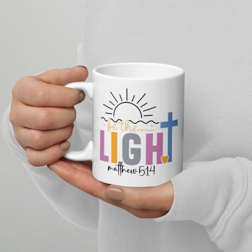 A person holds the "Be The Light Coffee Cup," featuring the text "be the LIGHT" and "Matthew 5:14," with a sun illustration above and a blue cross accompanied by a small yellow heart below. The person is dressed in a white long-sleeve top. christian gifts, religious gifts, gifts of faith, faith based gifts, faith inspired gifts