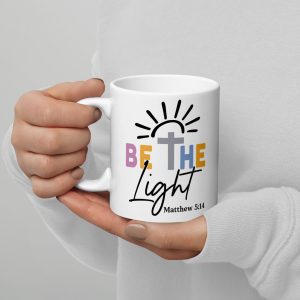 A person holding the "Be The Light Coffee Cup," a white mug adorned with the colorful phrase "BE THE Light," complemented by a cross and sun rays above, and featuring the verse "Matthew 5:14" printed below. The individual is wearing a white sweater and has pale nail polish. christian gifts, religious gifts, gifts of faith, faith based gifts, faith inspired gifts