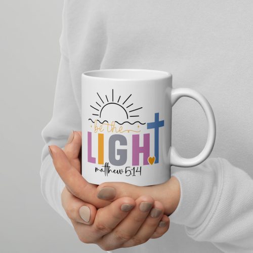 A person is holding the "Be The Light Coffee Cup," which features a design of a sun, a blue cross, and the phrase "be the LIGHT" alongside "Matthew 5:14" in decorative fonts on a white mug. The person's attire includes a light-colored sweater. christian gifts, religious gifts, gifts of faith, faith based gifts, faith inspired gifts