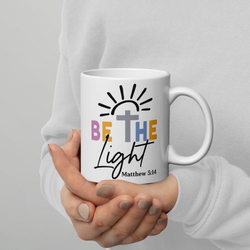 A person holds the Be The Light Coffee Cup featuring a colorful "Be the Light" phrase, along with a cross and sunburst graphic, and "Matthew 5:14" beneath. The person is dressed in a white sweater. christian gifts, religious gifts, gifts of faith, faith based gifts, faith inspired gifts