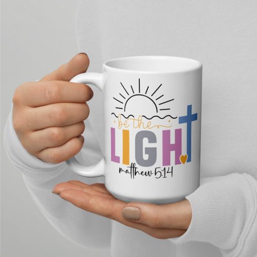 A person holds the Be The Light Coffee Cup, a white ceramic mug adorned with the phrase "Be the Light" in colorful letters, alongside "Matthew 5:14" and illustrations of a sun and cross. The individual is wearing a white long-sleeve shirt. christian gifts, religious gifts, gifts of faith, faith based gifts, faith inspired gifts