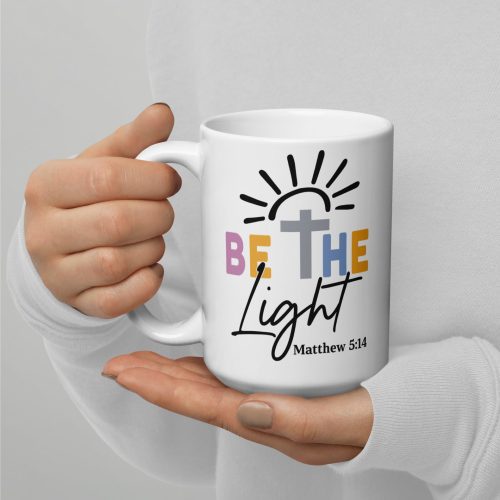 A person is holding the Be The Light Coffee Cup, which features a vibrant "Be the Light" design along with a sun and a cross. Below, there's a reference to Matthew 5:14. The individual is wearing a white long-sleeve shirt. christian gifts, religious gifts, gifts of faith, faith based gifts, faith inspired gifts