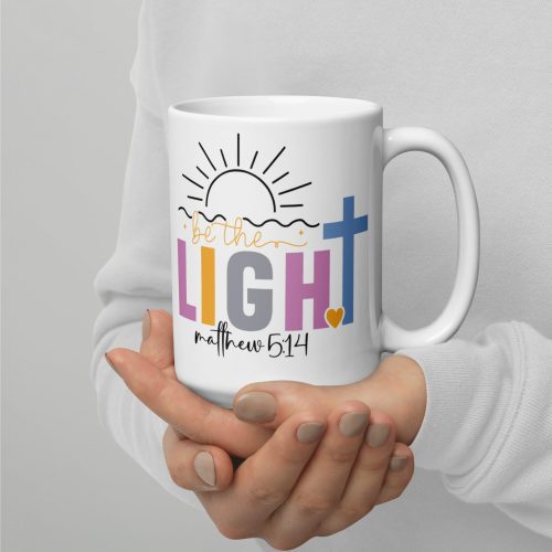 A person holds the "Be The Light Coffee Cup," which is a white mug featuring colorful text with the phrase "Be the Light" alongside the Bible reference "Matthew 5:14." The design incorporates a sun and a cross. The person is dressed in a light gray sweater. christian gifts, religious gifts, gifts of faith, faith based gifts, faith inspired gifts