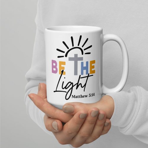 Against a plain white background, someone cradles the "Be The Light Coffee Cup," which displays the phrase "Be the Light" accompanied by a symbolic sun and cross design above and "Matthew 5:14" below. christian gifts, religious gifts, gifts of faith, faith based gifts, faith inspired gifts